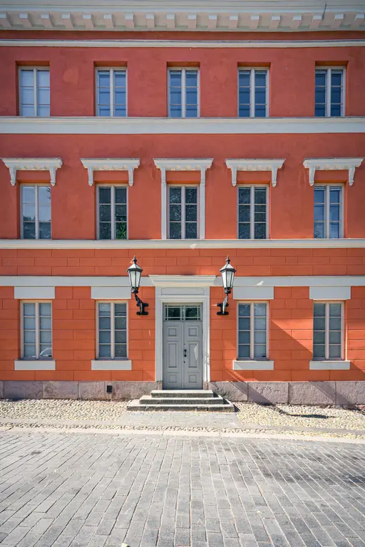 Fassade eines Hauses in warmem Orange mit zwei Lampen neben dem Eingang. Das Gebäude hat mehrere Fenster und eine graue Tür.