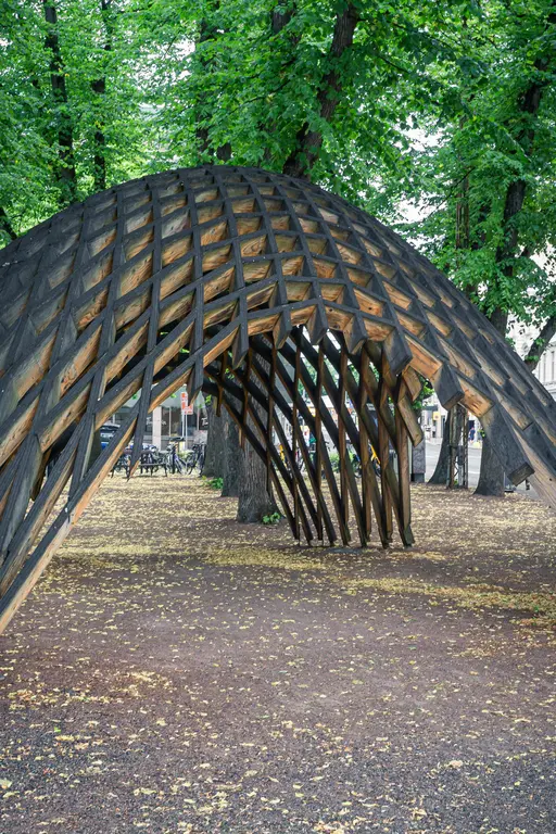 Holzskulptur in gewölbter Form mit offenem Design, umgeben von grünen Bäumen und herabfallenden Blättern auf dem Boden.