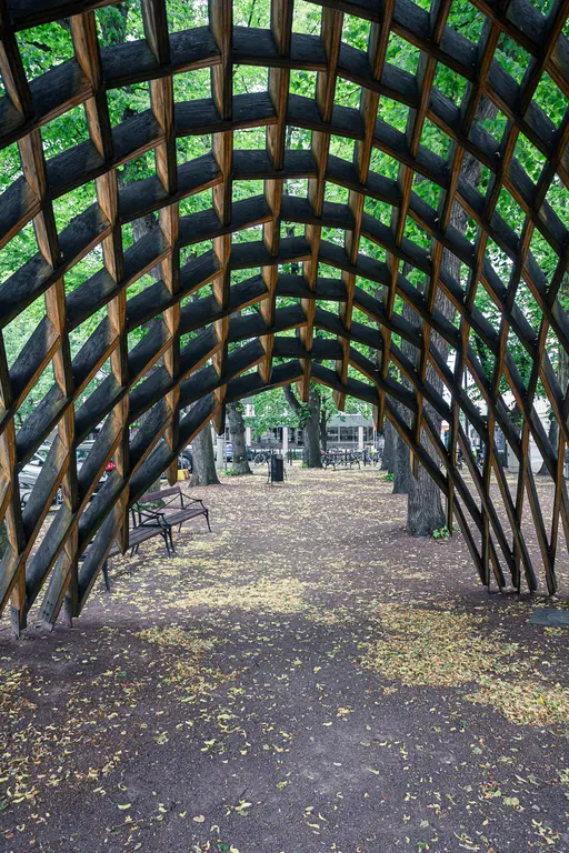 Holzlaubenstruktur mit gewölbtem Dach inmitten eines Parks, umgeben von grünen Bäumen und herabgefallenen Blättern auf dem Boden.
