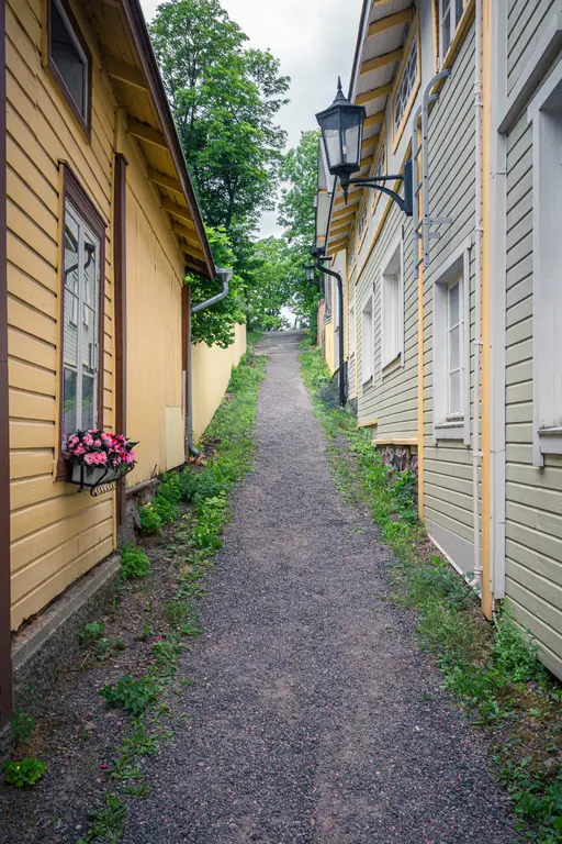 Pittoresker Weg zwischen zwei Holzgebäuden, flankiert von Grün und Blumen. Eine Straßenlaterne hängt an der Wand.