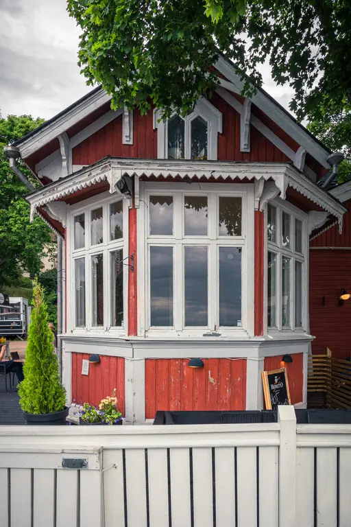 Rotes, hölzernes Gebäude mit großen Fenstern und dekorativen Elementen, umgeben von Bäumen und einer weißen Zaunfront.