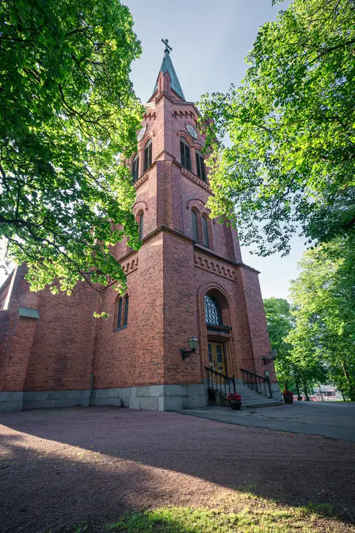 Ziegelsteinkirche mit hohem Turm, umgeben von grünen Bäumen. Die Fassade hat große Fenster und eine eingezogene Eingangstreppe.