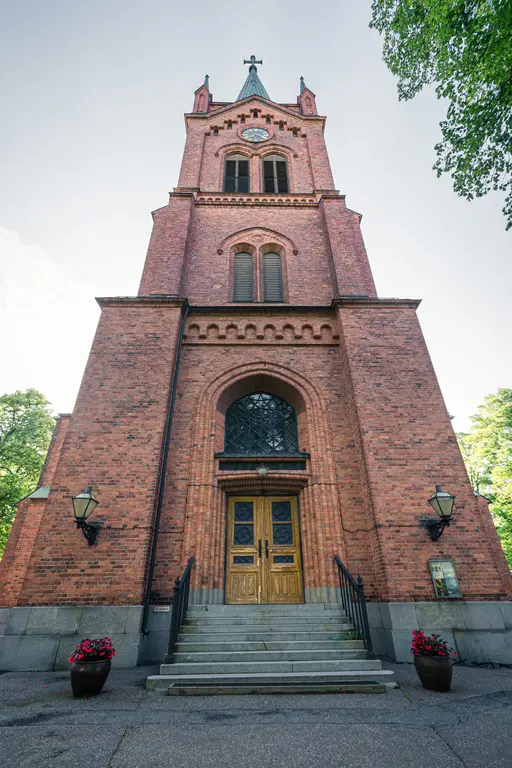 Roter Backsteinbau einer Kirche mit hohem Turm, großen Fenstern und einer auffälligen Holzportal-Tür. Stufen führen zur Eingangstür.