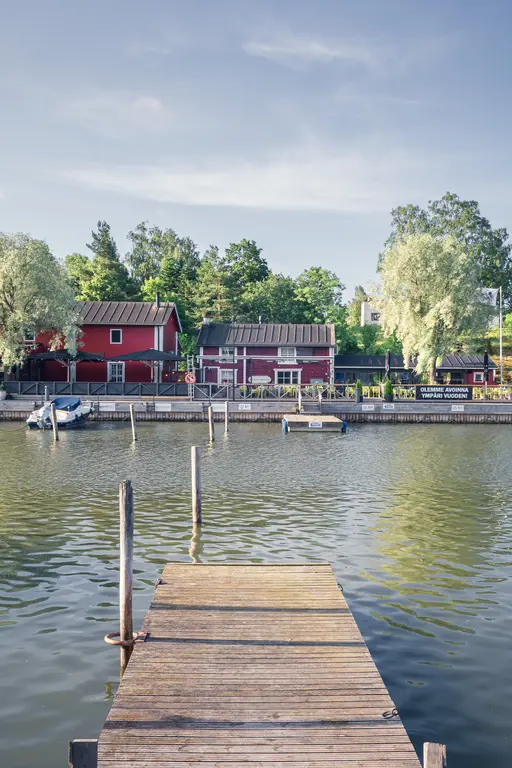 Holzsteg am Wasser mit rot gestrichenen Häusern im Hintergrund, umgeben von Bäumen und sanften Wellen.