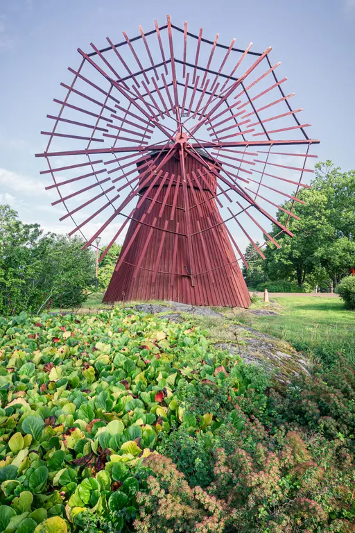 Rote Windmühle mit einem auffälligen, gitterartigen Flügelmechanismus, umgeben von grünen Pflanzen und Büschen.