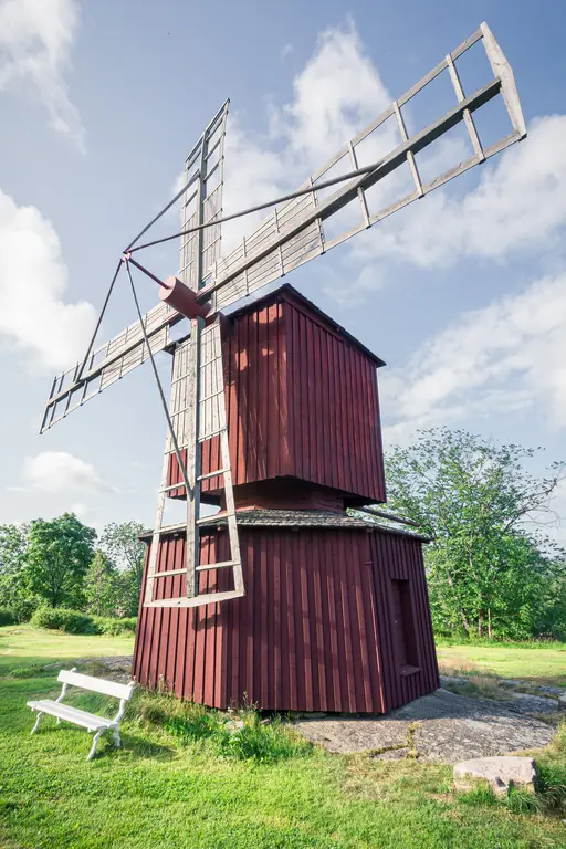 Rote Windmühle mit großen, schaufelartigen Flügeln steht auf einem grünen Rasen, umgeben von Bäumen und einem Banch.