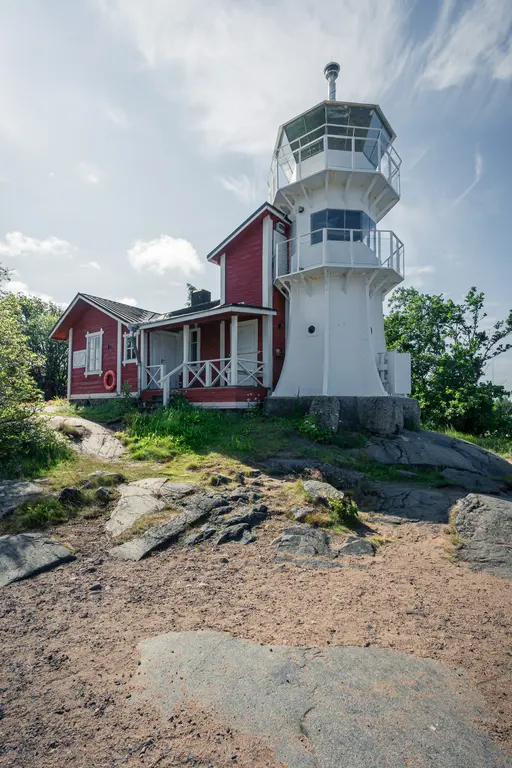 Roter Leuchtturm mit gläsernem Aussichtsturm, daneben ein kleines rotes Haus. Umgeben von felsigem Boden und grüner Vegetation.