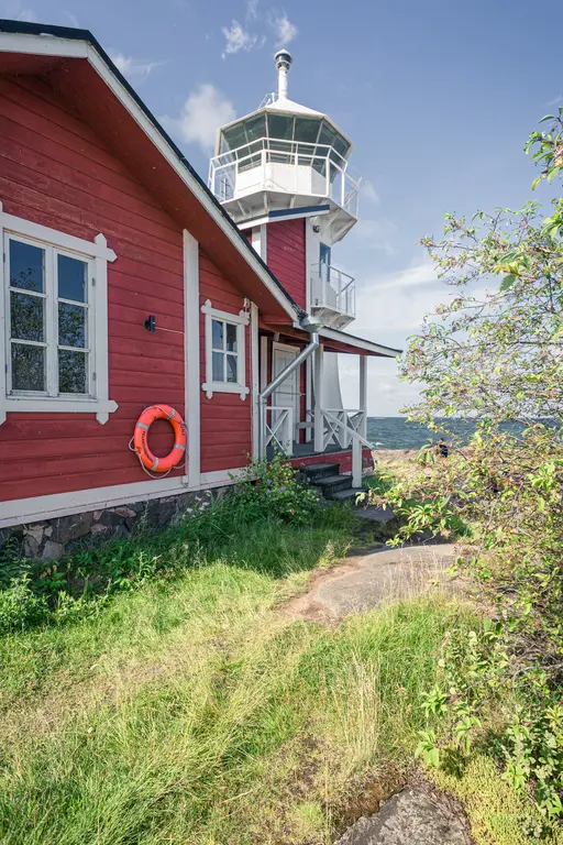 Leuchtturm aus rotem Holz neben einem Pfad, umgeben von grünem Gras und Sträuchern. Ein orangener Rettungsring hängt an der Wand.