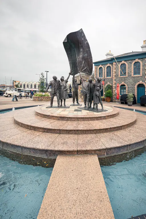 Bronzeskulptur einer Gruppe von Menschen, die gemeinsam eine Fahne halten, umgeben von einer kreisförmigen Steinplattform und Wasser.