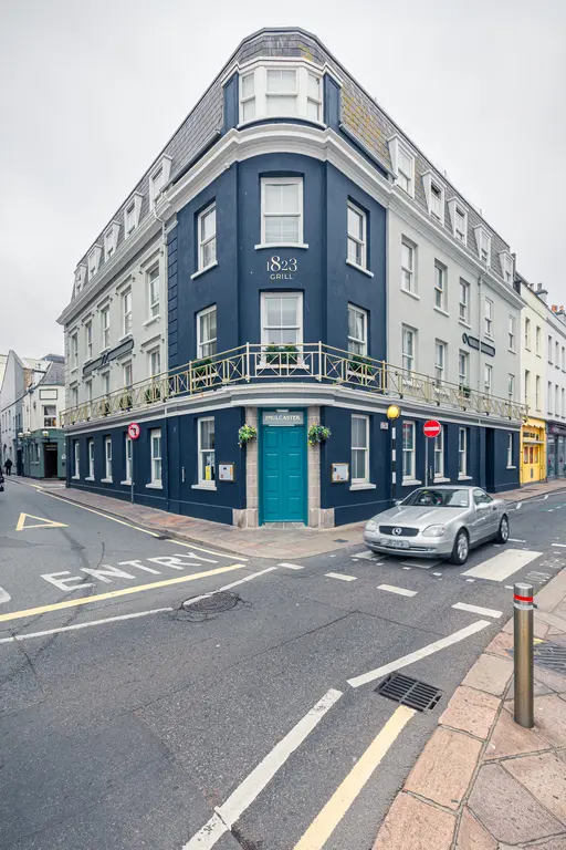 Eckgebäude mit graublauer Fassade und hellen Fensterrahmen, beschriftet mit „1823 Grill“. Eine Straße verläuft entlang der Front.