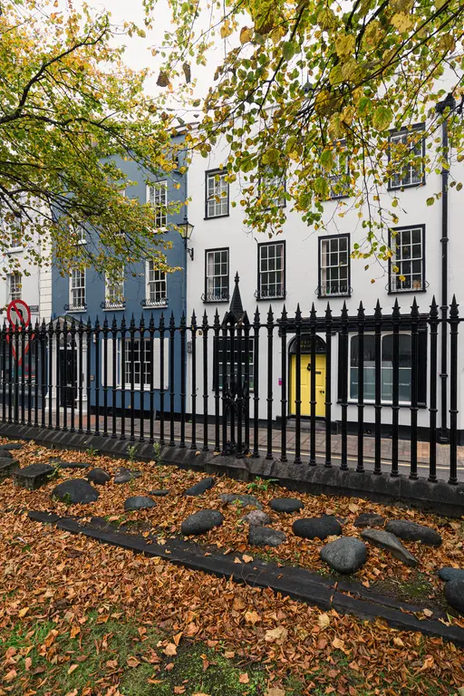Bunte Häuserfassaden in Blau und Weiß, mit einem gelben Eingang. Vor einem eisernen Zaun liegen orange-braune Herbstblätter und Steine.