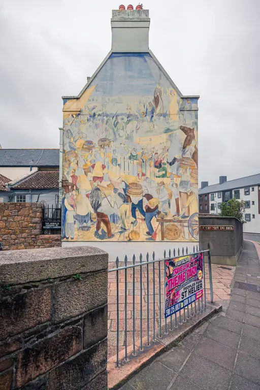 Wandgemälde an einem Gebäude, das eine lebhafte Marktszene darstellt. Im Vordergrund ist ein Zaun mit einem Plakat für einen Themenpark sichtbar.