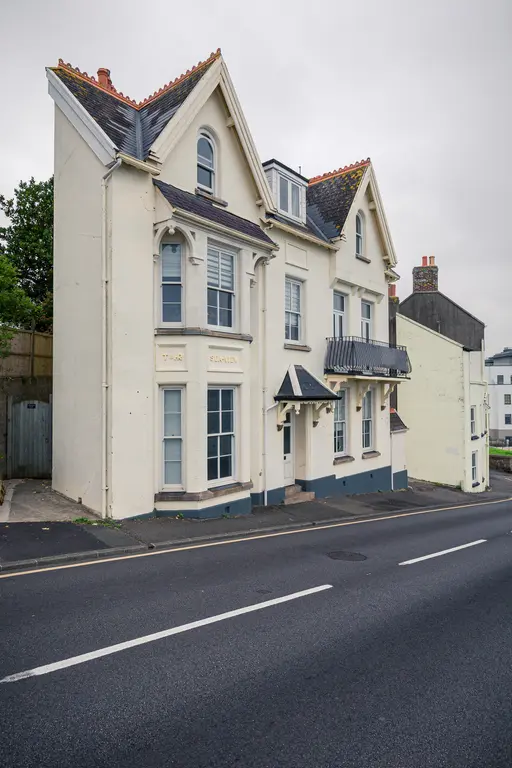 Dreistöckiges, helles Wohngebäude mit Satteldach und Fenstern im oberen Bereich. Eine kleine Veranda und eine gepflasterte Auffahrt sind sichtbar.