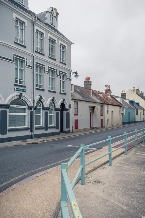 Städtische Straße mit grauem Gebäude und mehreren niedrigen Häusern. Eine blaue Umzäunung verläuft entlang des Gehwegs. Bewölkter Himmel.