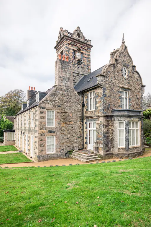 A historic stone building with a clock tower and detailed architecture, surrounded by green lawn and garden paths.