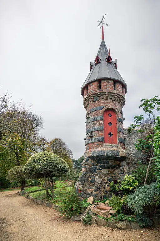A decorative stone tower with a red door and spire, surrounded by lush greenery and a walking path.