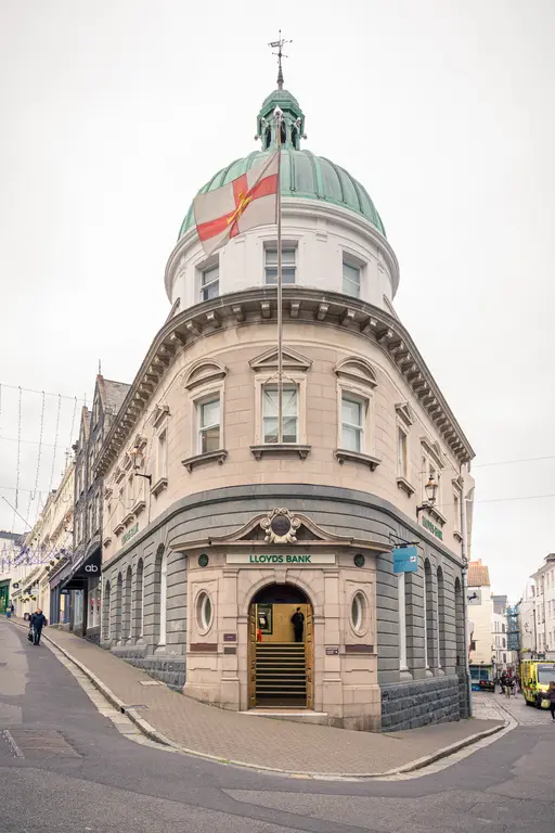 A historic bank building with a green dome, featuring an entrance and a flag on top, surrounded by sloped streets and shops.