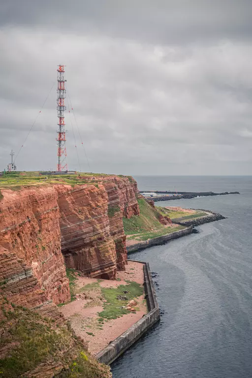 Eine steile, rote Klippe mit einer hohen Antenne und grünem Gras, die ins Wasser führt, an einem bewölkten Tag.