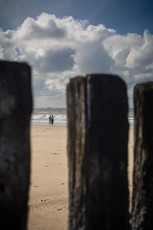 Ein Paar steht am Strand, umgeben von Holzpfählen; der Himmel ist bewölkt und das Meer ist sichtbar im Hintergrund.