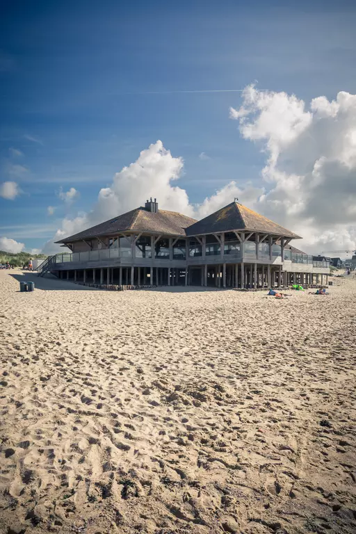 Ein modernes Strandgebäude mit Holzdetails steht auf Stelzen im Sand, umgeben von blauem Himmel und weißen Wolken.