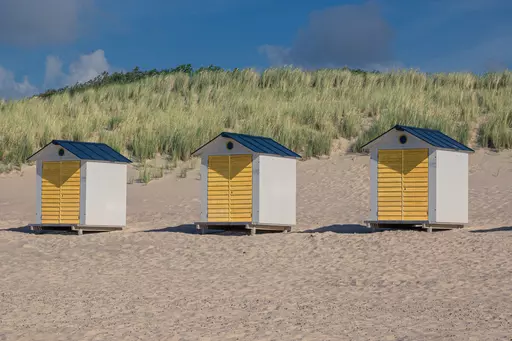 Drei Strandhäuschen mit gelben Türen stehen auf sandigem Boden, umgeben von grünen Dünen unter einem klaren Himmel.