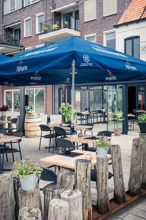 Tables with chairs and plants under a blue canopy. Wooden logs create a border. Inviting atmosphere.