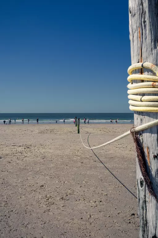 A post with rope in the foreground, with a wide sandy beach and people standing and moving near the water in the background.