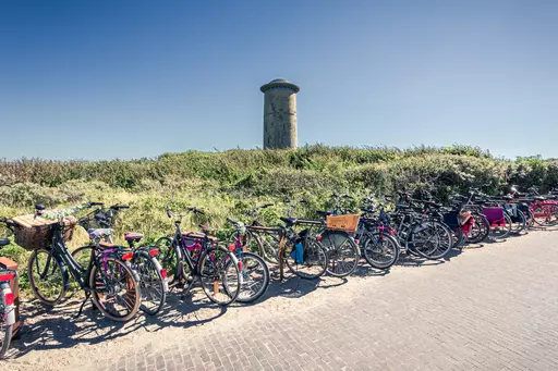 A line of bicycles is parked by a path, with a tall stone tower and lush greenery in the background.