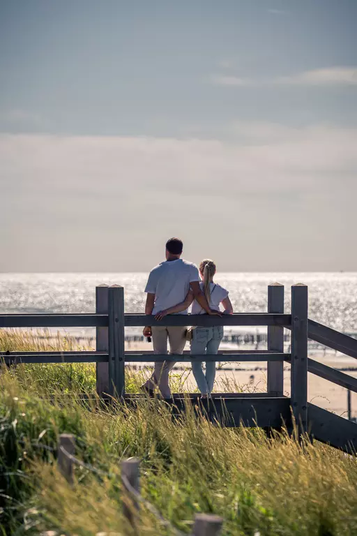 A couple stands by a wooden fence, gazing at the sparkling sea, surrounded by grass and sand.