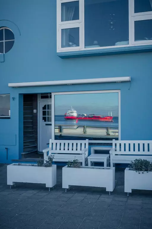 A blue wall with a large window reflects a red ship. White benches and planters stand in front of the window.