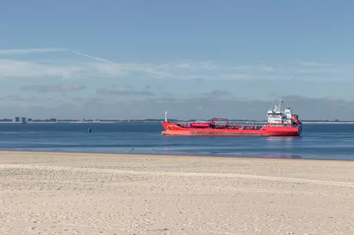 A red ship sails on calm water, with a sandy beach in the foreground and a lightly clouded sky in the background.