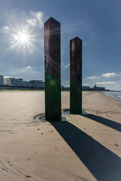 Two rusty pillars stand in the sand with the sun shining brightly above. The coastline is visible in the background.