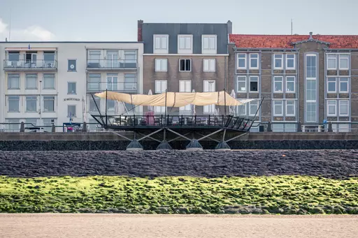 A modern café with a large sunshade sits on a waterfront promenade, surrounded by several multi-story buildings.