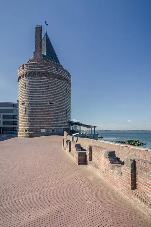 A round brick tower with a pointed roof stands by the water. A cobblestone walkway runs along the shore.