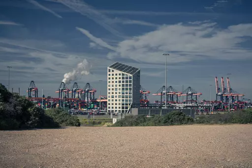 A modern, tilted building stands in the foreground with cranes and containers visible in the background.