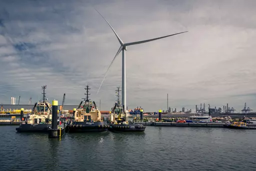 Three tugboats rest in the water, surrounded by a harbor landscape and a large wind turbine in the background.