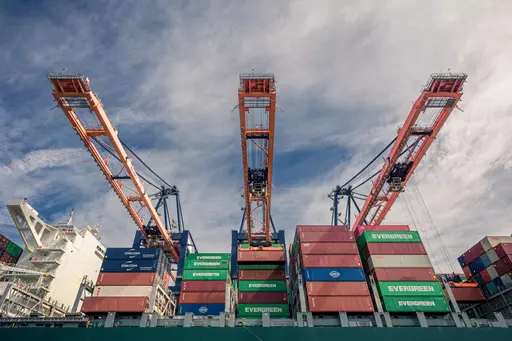 Three large cranes lift containers from a freighter loaded with colorful containers under a cloudy sky.