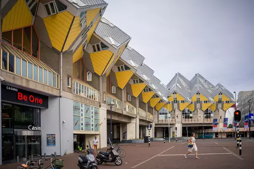 Colorful, cubic buildings with yellow roofs dominate the scene, and a woman is walking by in the foreground.