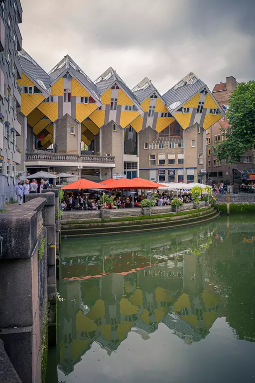 Colorful houses with trapezoidal roofs are reflected in the water, surrounded by a lively restaurant area under orange umbrellas.