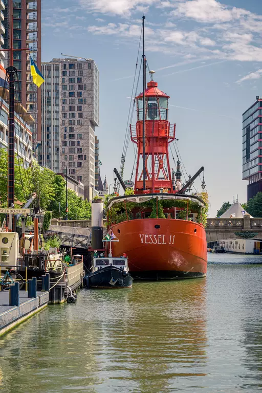 A red ship named Vessel 11 is docked along a canal, surrounded by modern buildings and lush greenery.