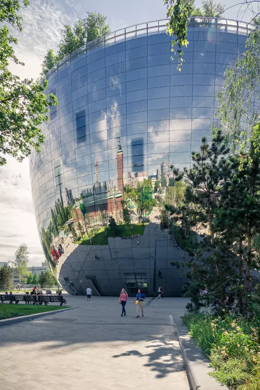 A modern, mirrored building facade reflects the surrounding landscape and people on a walkway, with green trees visible.