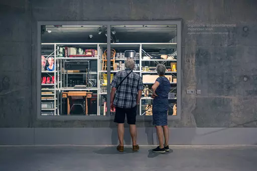 Two people are viewing a lit shelf filled with various items through a window.