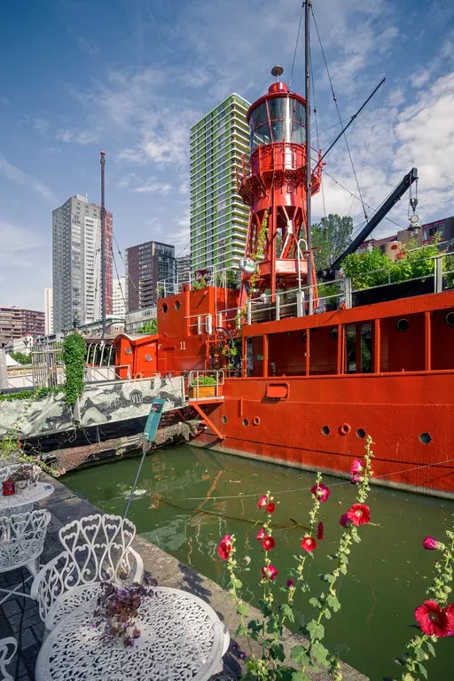 A red ship lantern stands next to an old red ship by a water canal, surrounded by modern buildings and blooming roses.