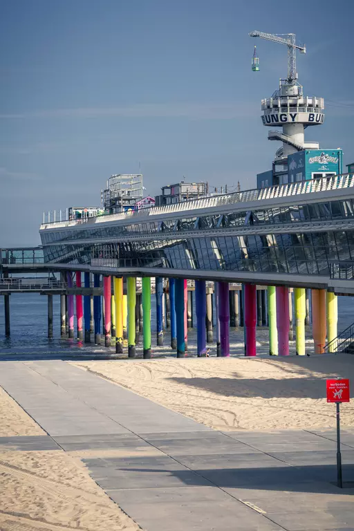 Colorful pillars support a walkway over the water. A sandy beach and a warning sign are visible in the foreground.