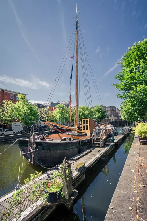 A historic boat rests by a canal surrounded by trees and modern buildings under a clear blue sky.