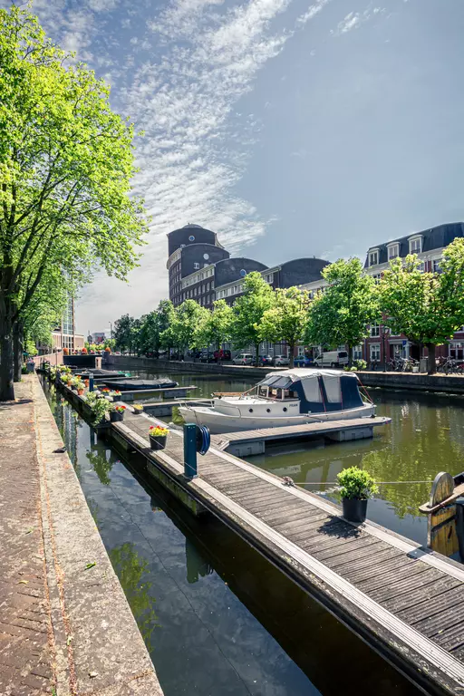 Green trees line a canal with several boats. The quay features flower boxes and buildings in the background.