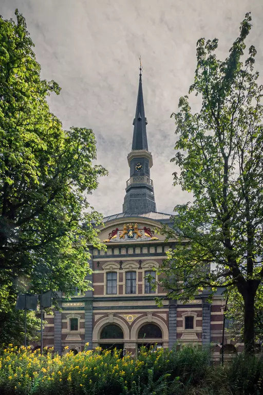 A view of a historic building with a tall pointed tower and colorful ornaments, surrounded by trees and yellow flowers.