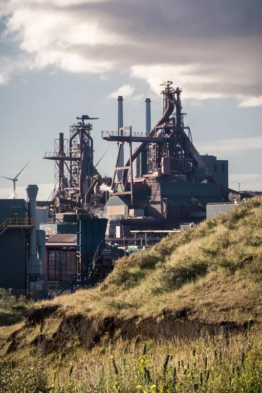 Industrial plant with tall towers and pipes, surrounded by grass and hills under a cloudy sky.