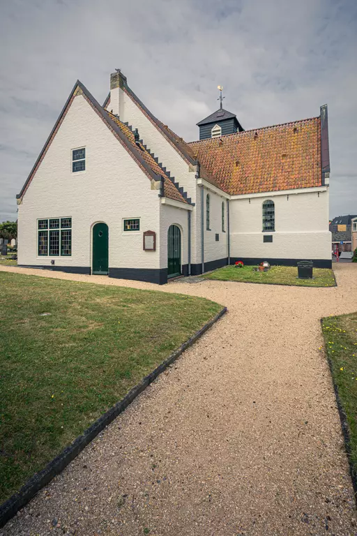 Eine weiße Kirche mit einem rot gefliesten Dach, umgeben von Rasen und Kieswegen, unter einem bewölkten Himmel.