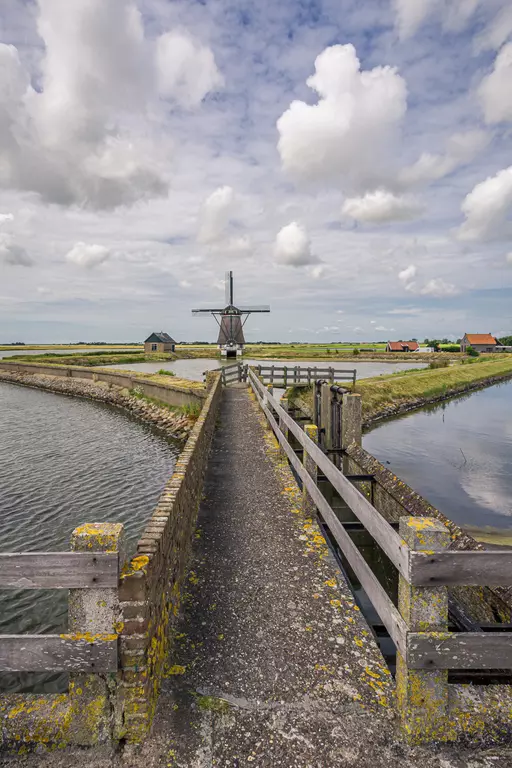 Eine Steinbrücke führt zu einer Windmühle, umgeben von Wasser und einem grasbewachsenen Ufer unter einem bewölkten Himmel.